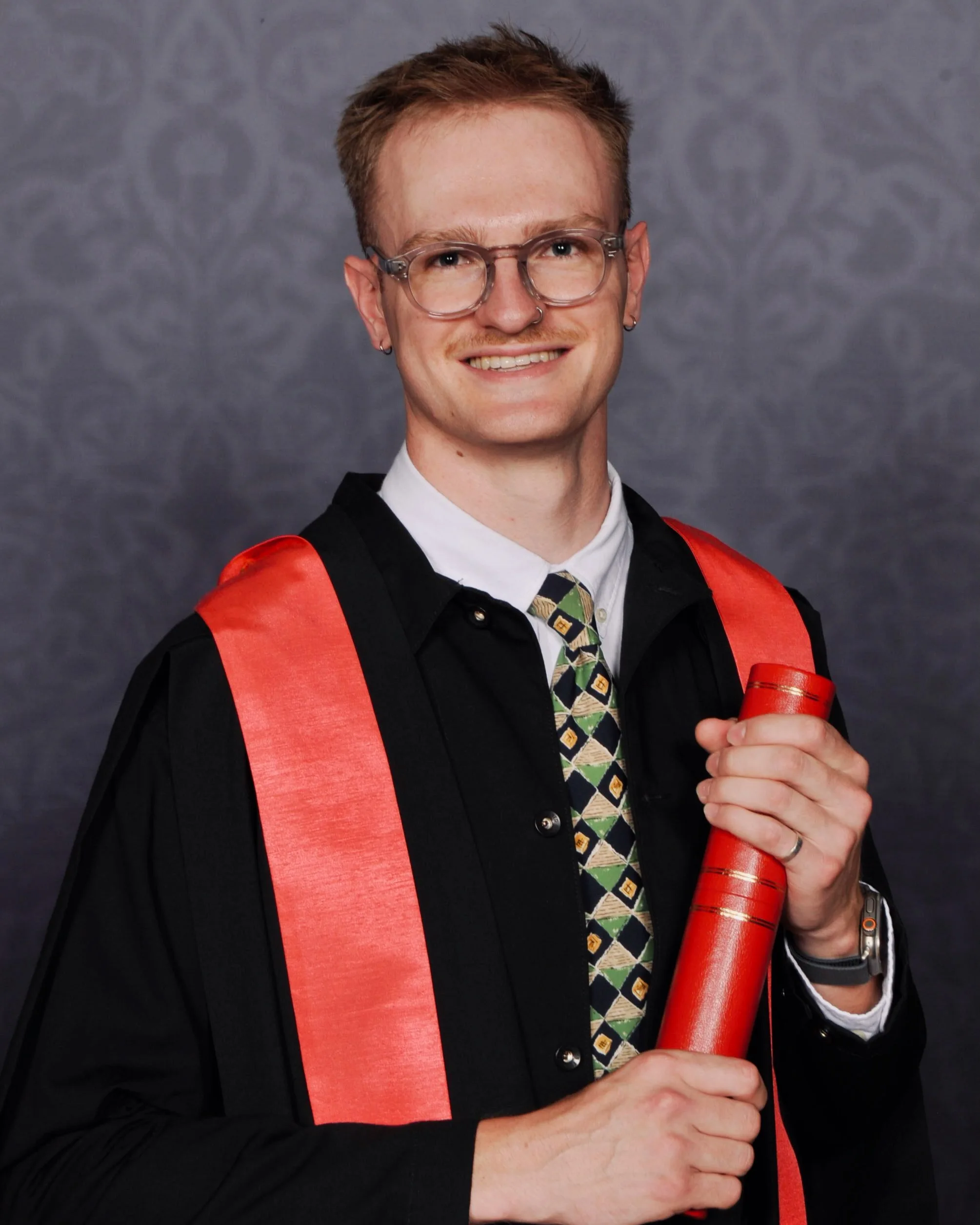 Noah sat smiling wearing his Kings College London graduation gown and holding his degree scroll