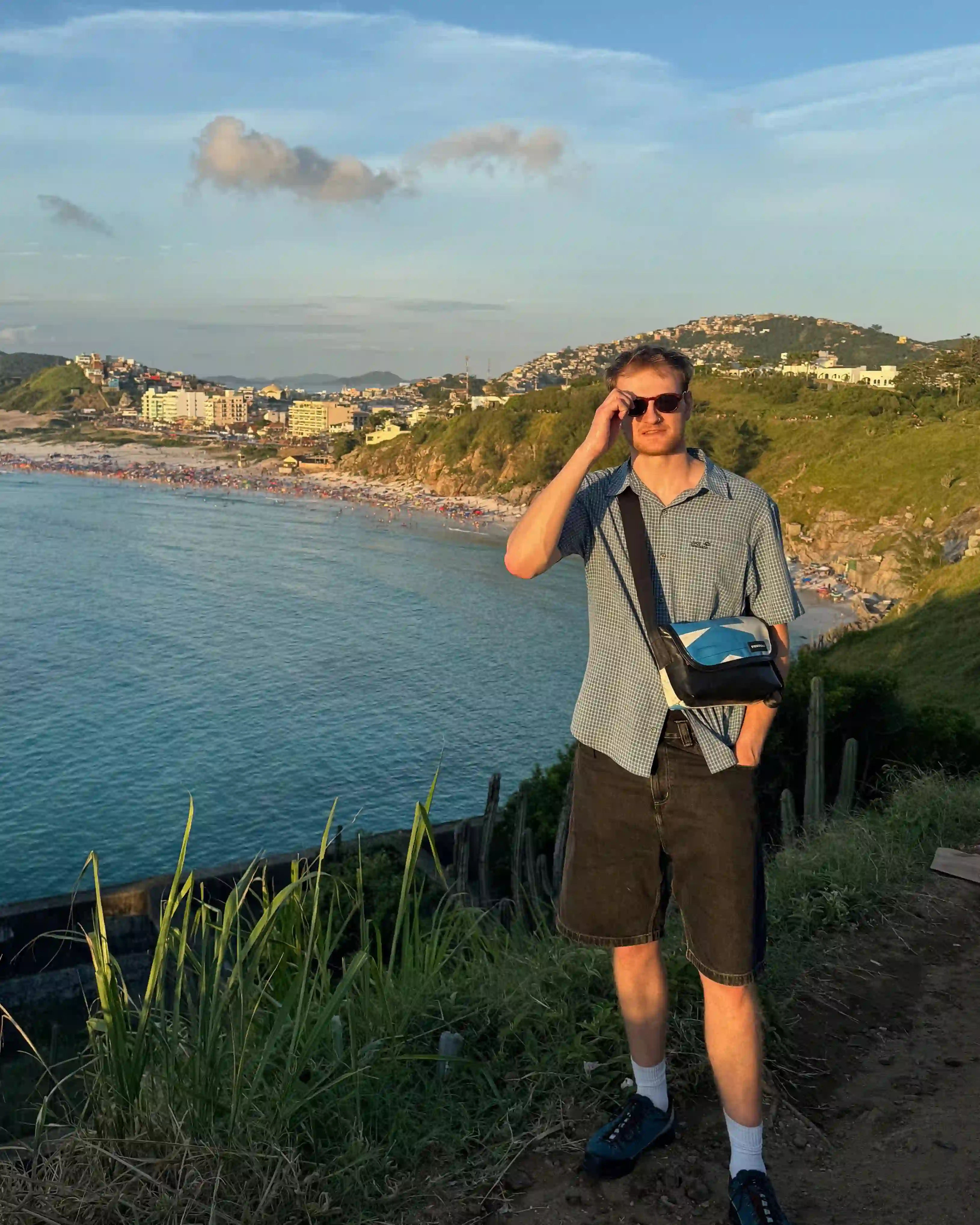 A landscape with a sunset over a beach cove in the background and Noah stood in the foreground adjusting his sunglasses